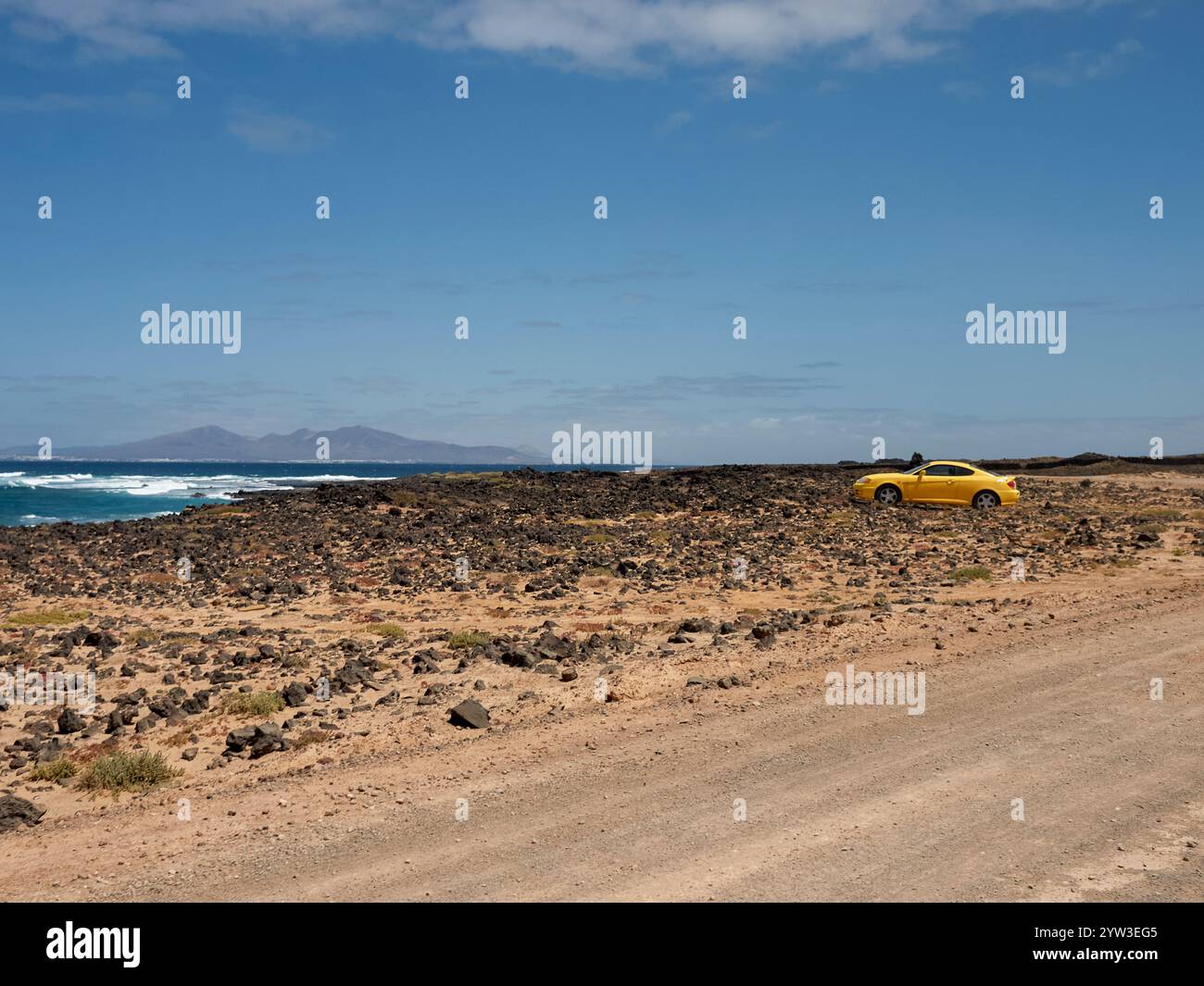 Gelbes Auto parkt an einer unbefestigten Straße in der Nähe der felsigen Küste mit Wellen und klarem blauem Himmel, Fuerteventura, Spanien Stockfoto