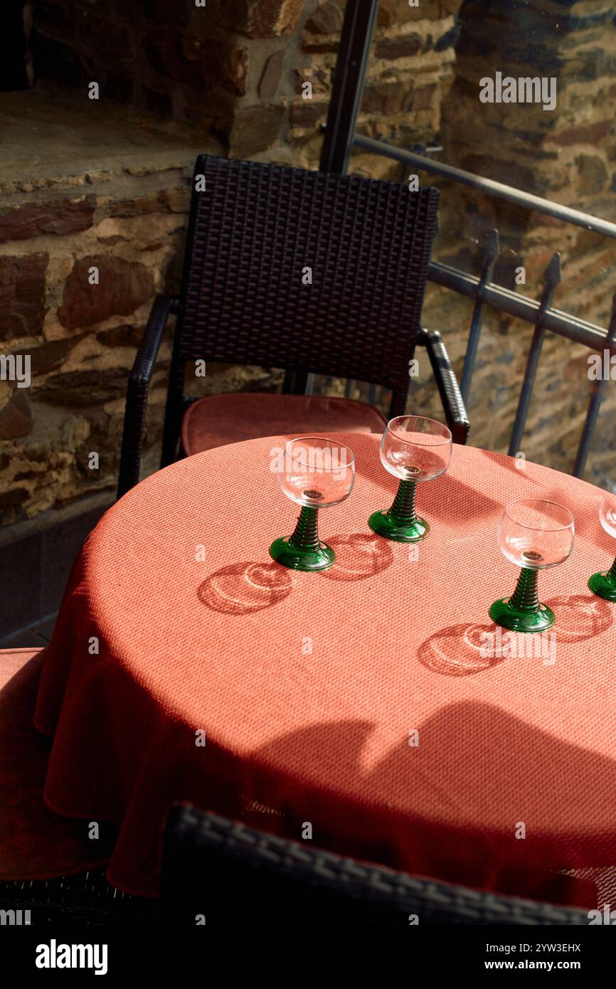 Sonnenlicht wirft Schatten auf einen Tisch im Freien, der mit grünen Gläsern besetzt ist, in einem malerischen Restaurant an der Mosel Stockfoto