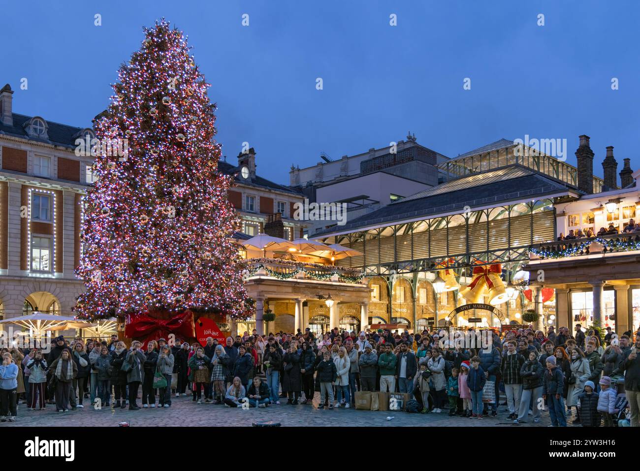 London, Vereinigtes Königreich - 2. Dezember 2024: Covent Garden Shopping Centre, in dem Menschen Straßenkünstler an einem großen beleuchteten Weihnachtsbaum beobachten Stockfoto