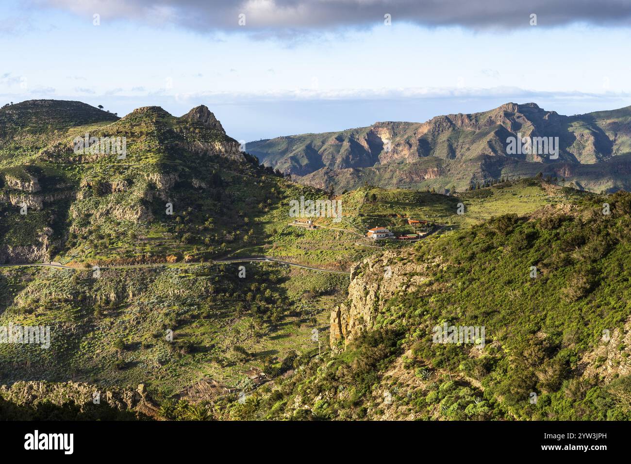 Landschaft im Südosten von La Gomera. Einige Häuser im Dorf Vegaipala. Berghänge mit Palmen und Agaven, einer Straße, Sonne und Wolken. Stockfoto