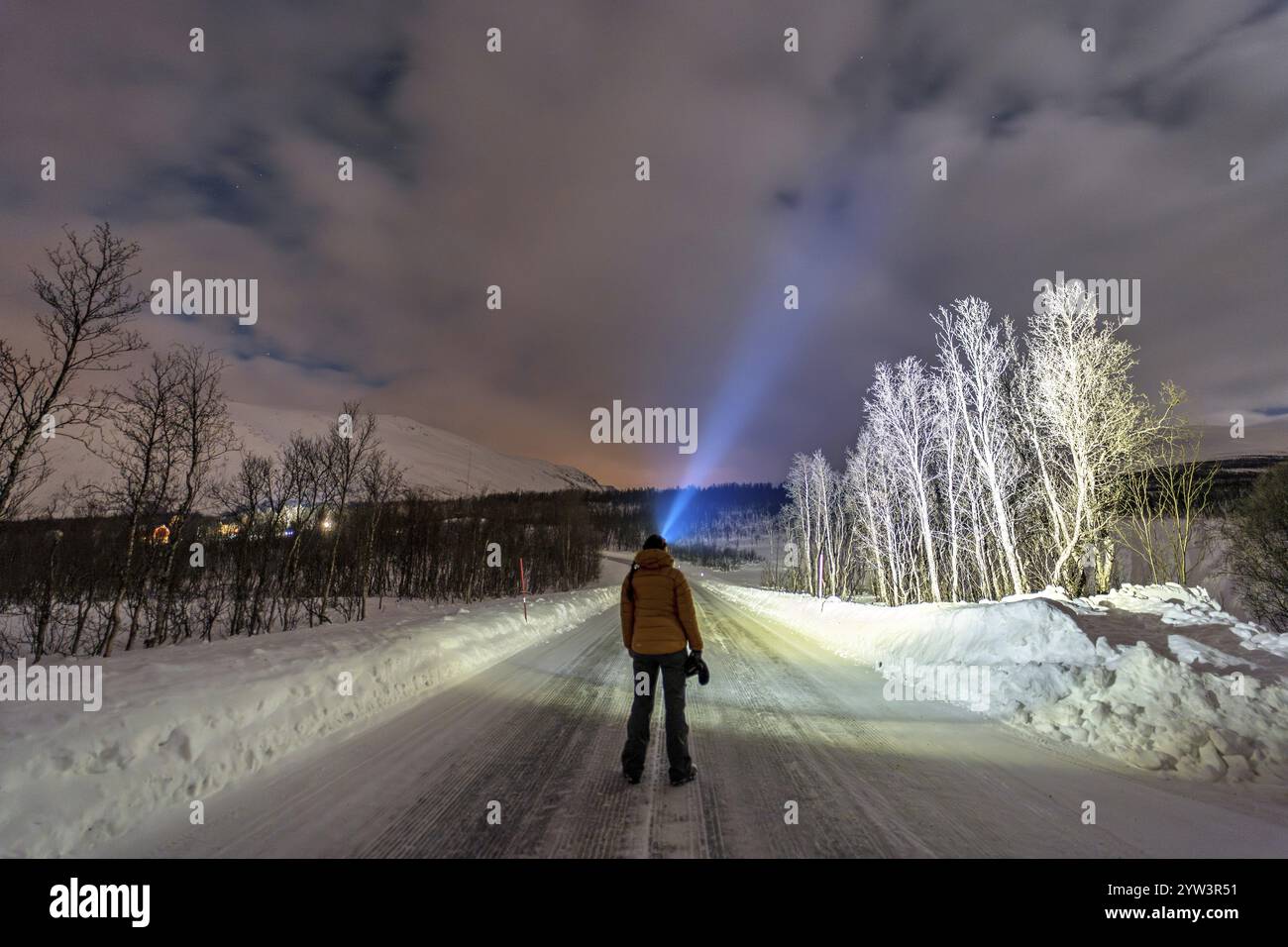 Eine Frau in Winterkleidung steht nachts auf einer verschneiten Straße und strahlt eine Stirnlampe in den Himmel, Tromso, Troms, Norwegen, Europa Stockfoto