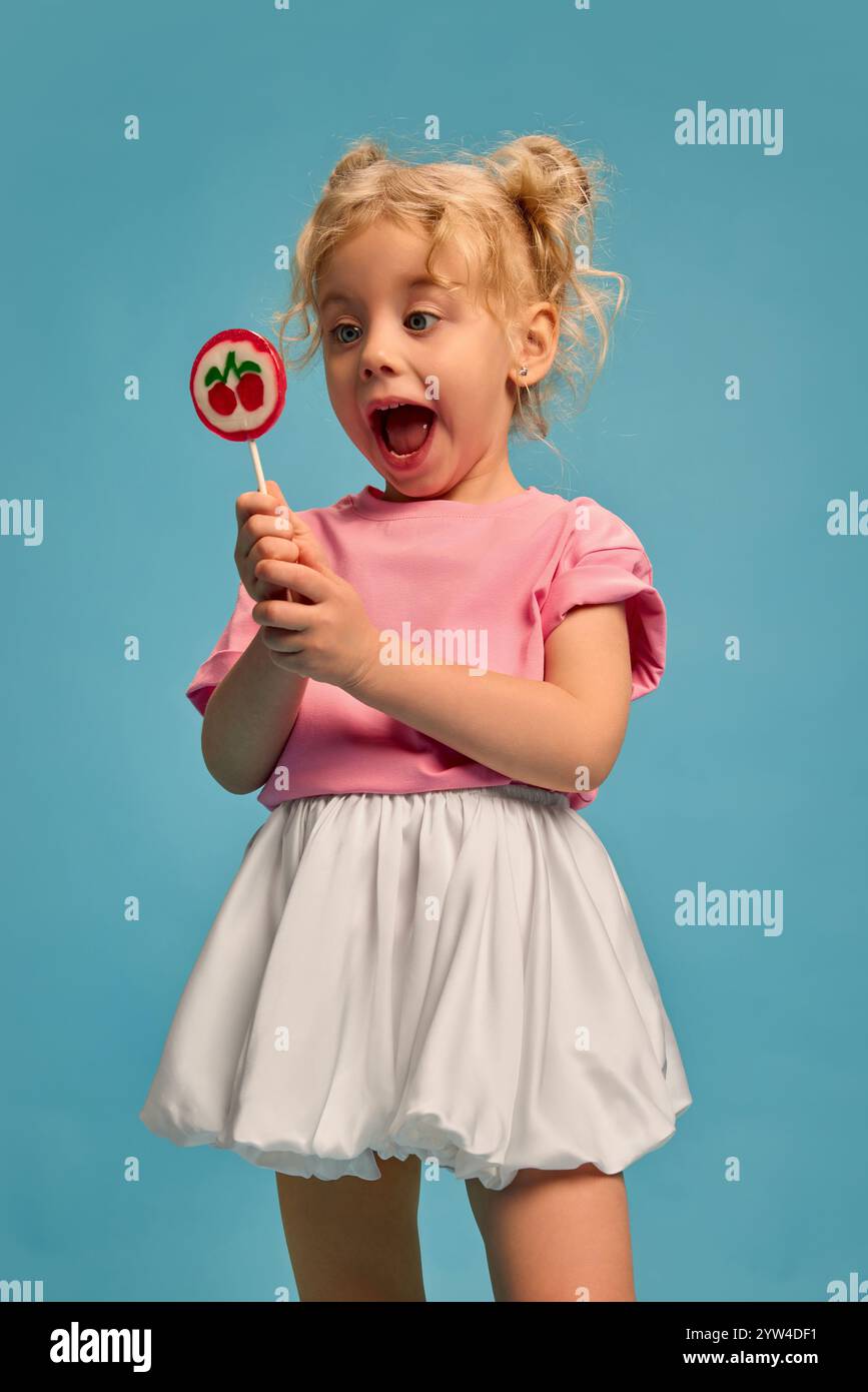 Porträt eines glücklichen, emotionalen kleinen Mädchens in rosa Hemd und weißem Rock, der Lutscher mit weit geöffnetem Mund nach der Impfung hält. Stockfoto