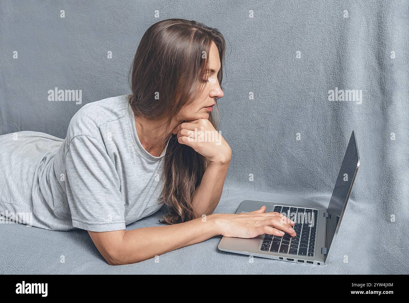 Seitenansicht der Hände der Frau mit Laptop beim Liegen auf dem Sofa. Langes Haar, graues T-Shirt, konzentrierter Look Stockfoto