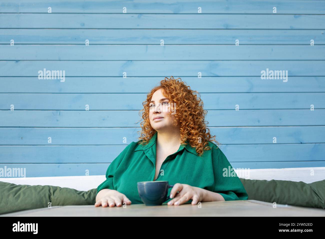 Schöne Frau in Übergröße mit rotem Haar, die eine Tasse Kaffee genießt. Kurviges weibliches Model sitzt am Cafe-Tisch in grünem Kleid gegen helles buntes blaues W Stockfoto