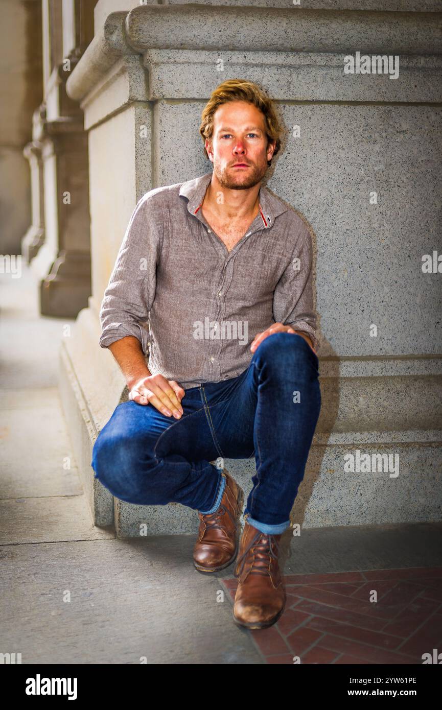 Eine Person mit kontemplativem Ausdruck hockt sich neben einer Steinstruktur in lässigem Hemd und Jeans. Warmes Licht wirft sanfte Schatten, Stockfoto