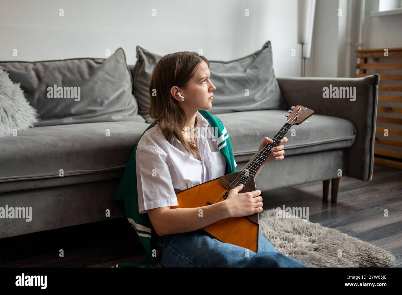 Eine aufmerksame Frau mit Ohrstöpseln hört Musik, während sie Balalaika-Saiten auf dem Boden im Wohnzimmer spielt Stockfoto