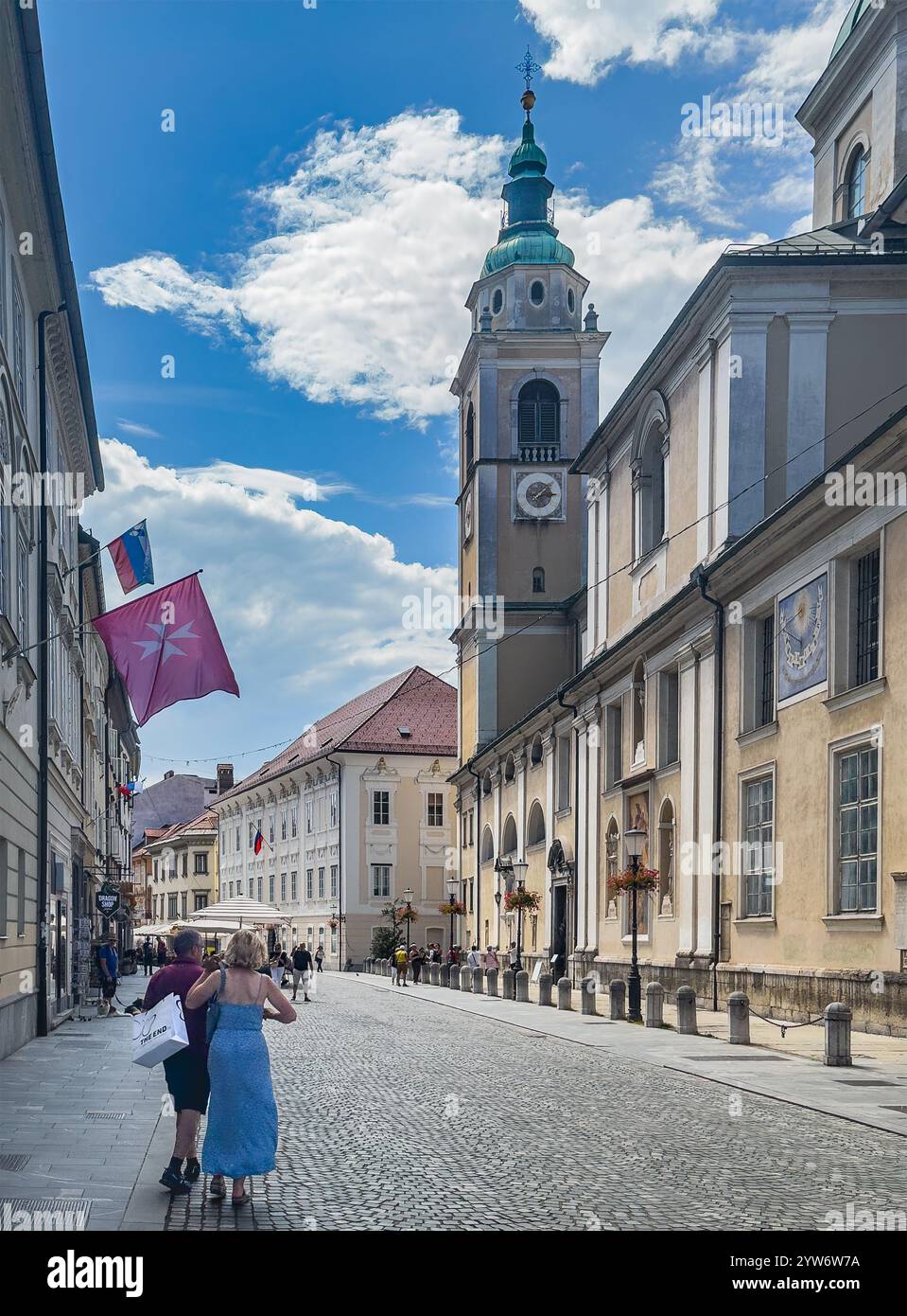 Ljubljana, Slowenien - 28. Juni 2024: Nikolaus-Kathedrale mit Uhrenturm von der Einkaufsstraße Ciril-Metodov unter blauer Wolkenlandschaft. Pedest Stockfoto
