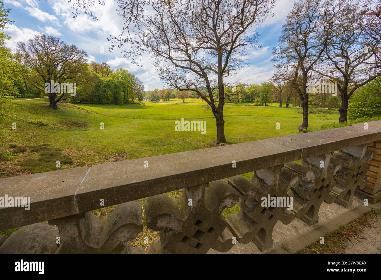 Wunderschöne Landschaft des großen grünen Parks mit grünen Lichtungen, Bäume am bewölkten sonnigen Morgen gesehen von einer kleinen Brücke mit Betonzaun im Park Stockfoto