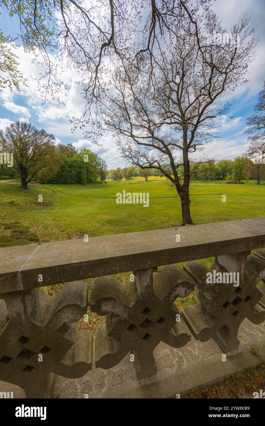 Wunderschöne Landschaft des großen grünen Parks mit grünen Lichtungen, Bäume am bewölkten sonnigen Morgen gesehen von einer kleinen Brücke mit Betonzaun im Park Stockfoto