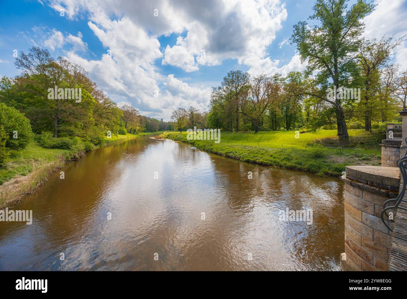 Wunderschöne Landschaft von einer kleinen Brücke über einen langen Fluss im Park voller grüner Lichtungen, Bäume und Büsche am sonnigen bewölkten Morgen Stockfoto