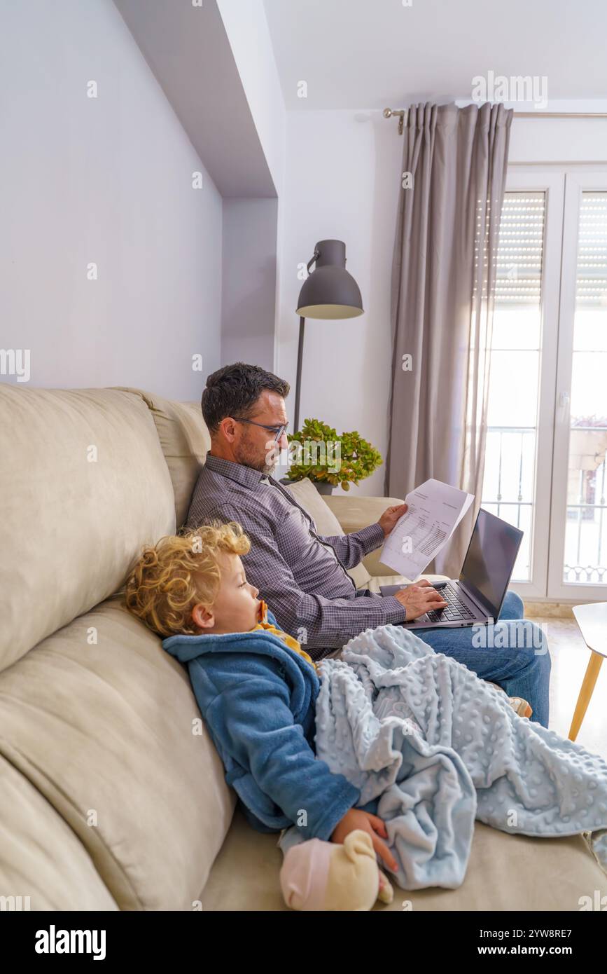 Vater Multitasking zu Hause, arbeitet aus der Ferne, während er sich um sein krankes Kleinkind kümmert, und hebt die Balance zwischen Beruf und Familie hervor Stockfoto