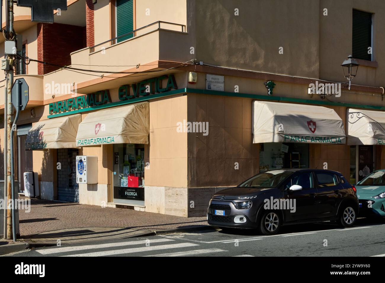 Savona. Italia - 10. Dezember 2024: Apotheke an einer sonnigen Straßenecke in Savona, Italien, mit geparkten Autos und einer Fußgängerüberfahrt davor Stockfoto