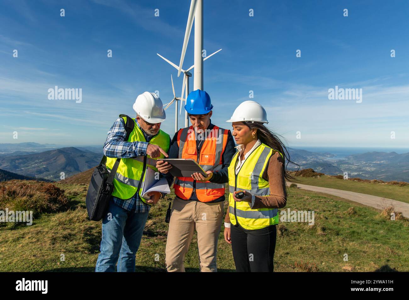 Drei multiethnische Ingenieure, die Sicherheitswesten und Helme tragen, verwenden ein digitales Tablet und diskutieren neben Windturbinen auf einem Hügel mit Blick Stockfoto