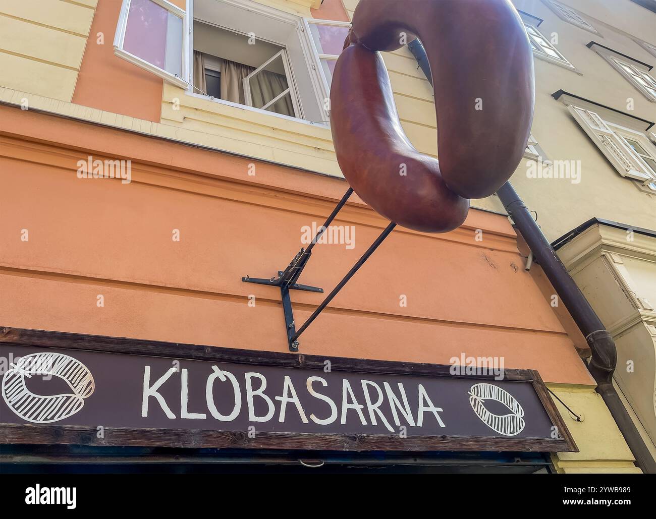 Ljubljana, Slowenien - 28. Juni 2024: Klobasarna-Wurstladen-Emblem, dreidimensionales Kunstwerk von 2 Blutwürsten vor blauer Wolkenlandschaft in Ciril-Metodo Stockfoto