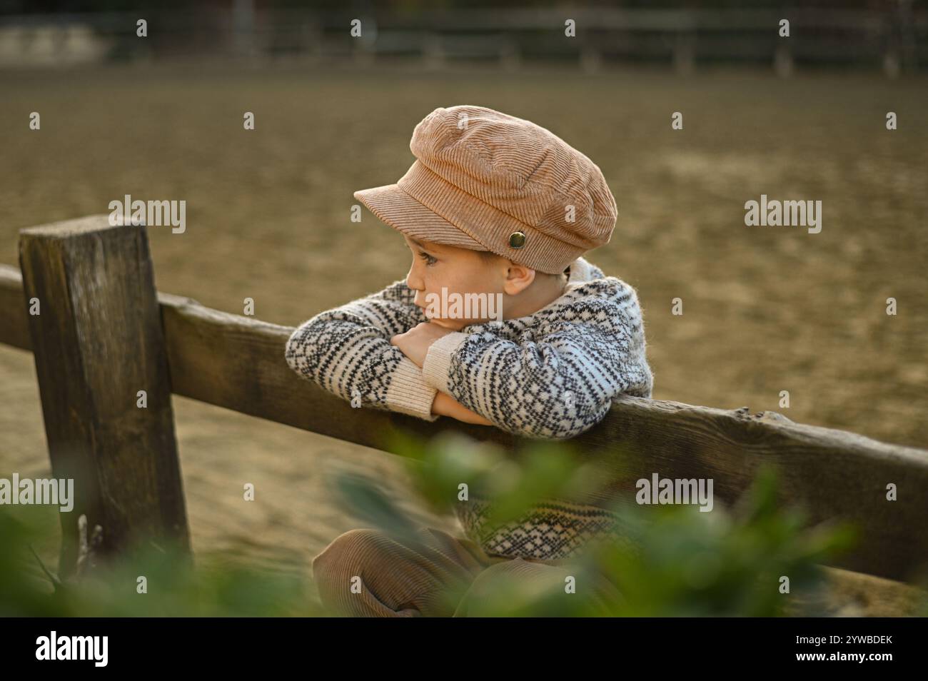 Junge in einem alten Hut und Pullover auf dem Zaun einer Reitarena Stockfoto