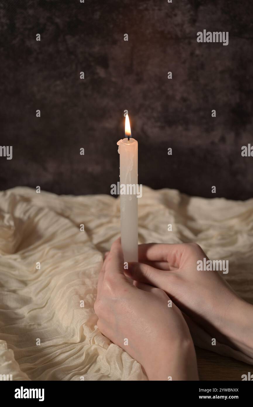 Eine weiße Kerze in Frauenhänden auf dunkelgrauem Hintergrund, beigefarbenes Tuch Stockfoto
