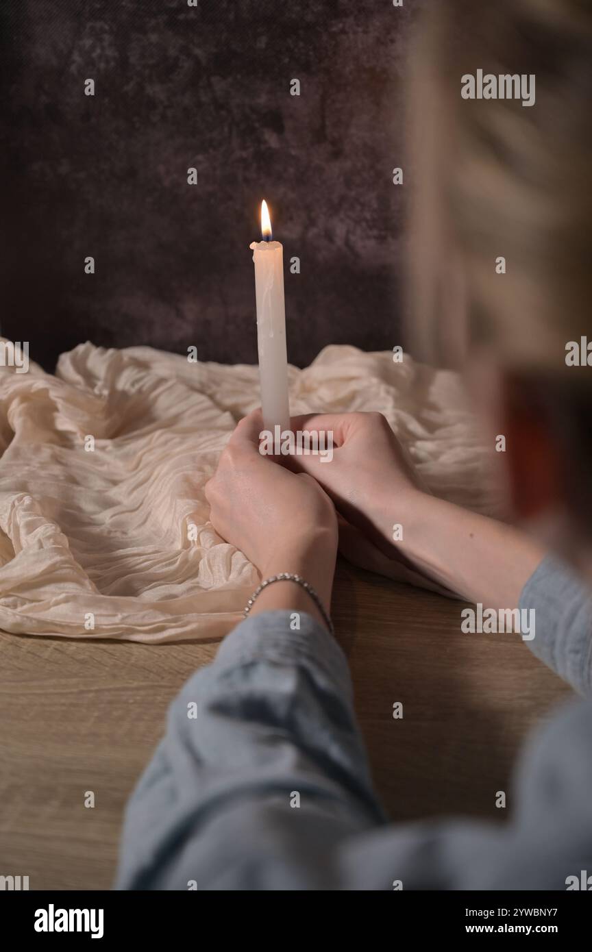 Eine weiß beleuchtete Kerze in betenden Frauenhänden auf dunkelgrauem Hintergrund, beigefarbenes Tuch auf Holztisch Stockfoto