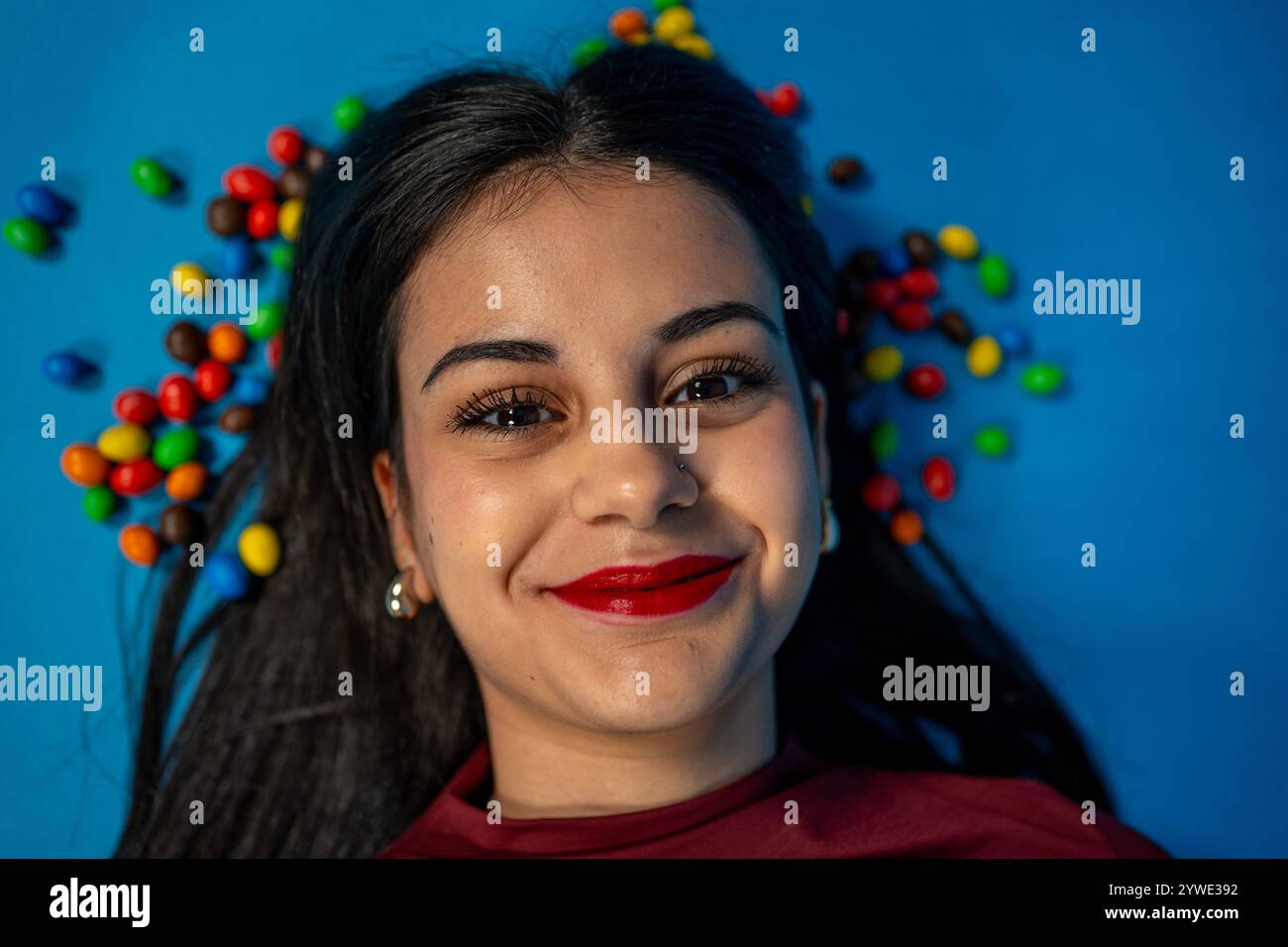 Porträt einer fröhlichen jungen Frau mit rotem Lippenstift auf einem leuchtend blauen Hintergrund, umgeben von bunten Bonbons, die Glück und Enjo ausdrücken Stockfoto