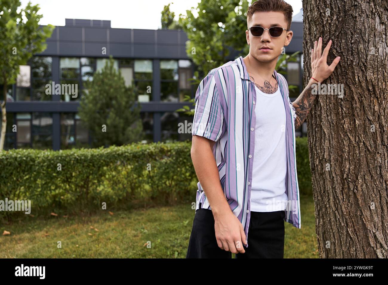 Ein junger Mann steht selbstbewusst an einem Baum in einem lässigen Outfit. Stockfoto
