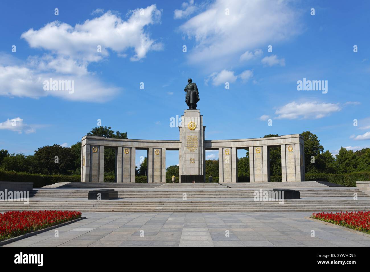 Ein großes Denkmal mit einer Statue auf einer Plattform, umgeben von Säulen und Blumenbeeten unter einem blauen Himmel mit weißen Wolken, sowjetisches Denkmal, Berlin, Tierga Stockfoto