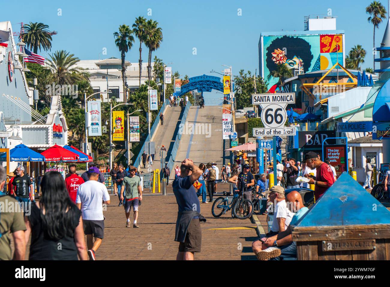 Touristen wandern entlang des Santa Monica Pier, der das Ende der Route 66 mit Geschäften, Ständen und Restaurants markiert. Blauer, sonniger Himmel Stockfoto