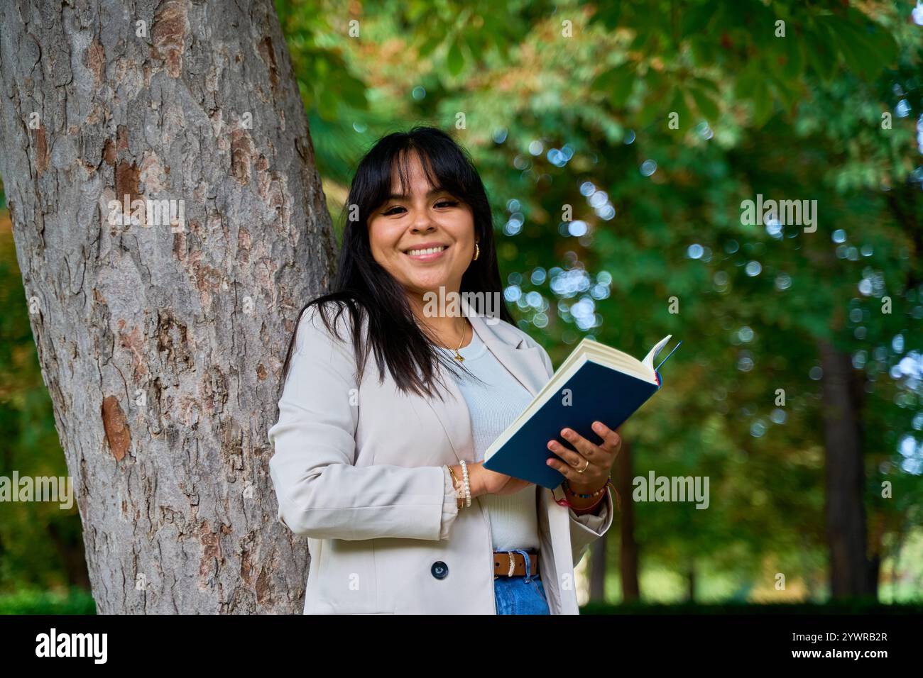 Latina liest ein Buch, während sie sich an einen Baum im Park lehnt Stockfoto