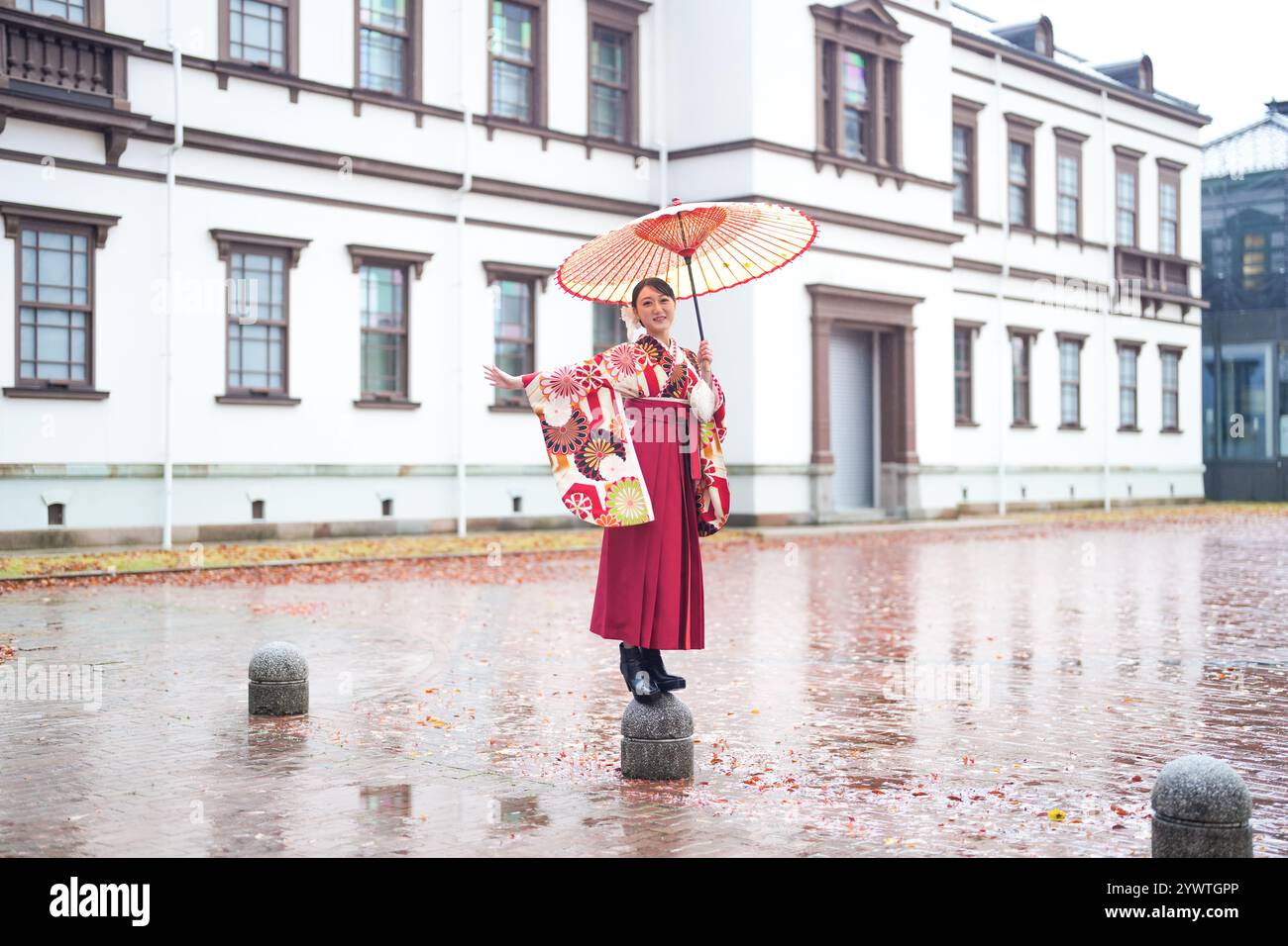 Eine Japanerin im Alter von 20 Jahren, die einen roten Kimono (Hakama) trägt, ein Grundnahrungsmittel japanischer Universitätsabsolventen, posiert im Regen mit einem roten japanischen Regenschirm Stockfoto