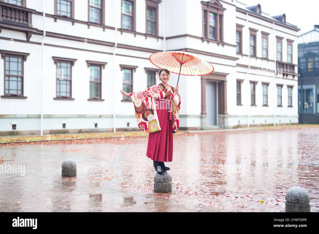 Eine Japanerin im Alter von 20 Jahren, die einen roten Kimono (Hakama) trägt, ein Grundnahrungsmittel japanischer Universitätsabsolventen, posiert im Regen mit einem roten japanischen Regenschirm Stockfoto