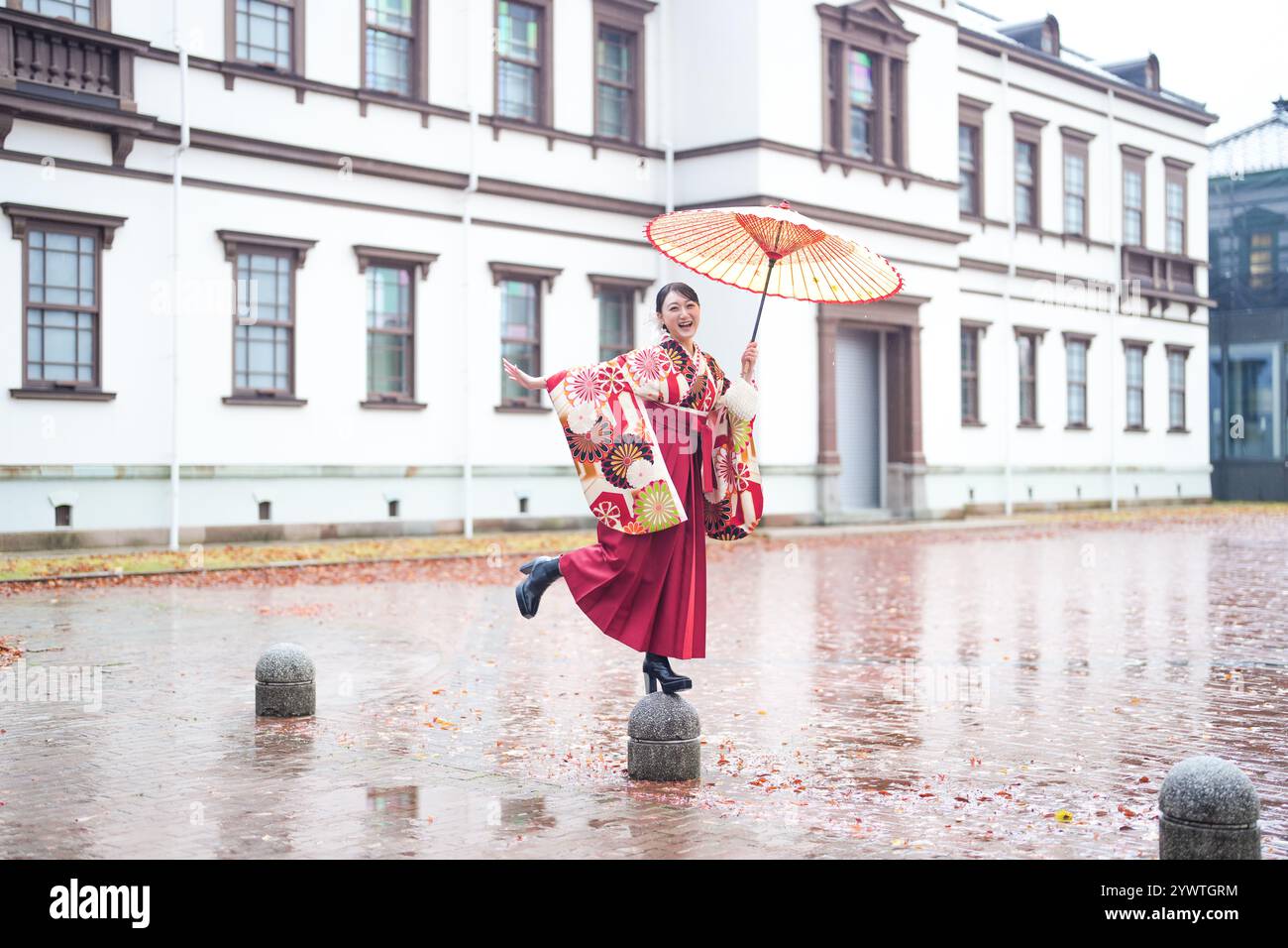 Eine Japanerin im Alter von 20 Jahren, die einen roten Kimono (Hakama) trägt, ein Grundnahrungsmittel japanischer Universitätsabsolventen, posiert im Regen mit einem roten japanischen Regenschirm Stockfoto