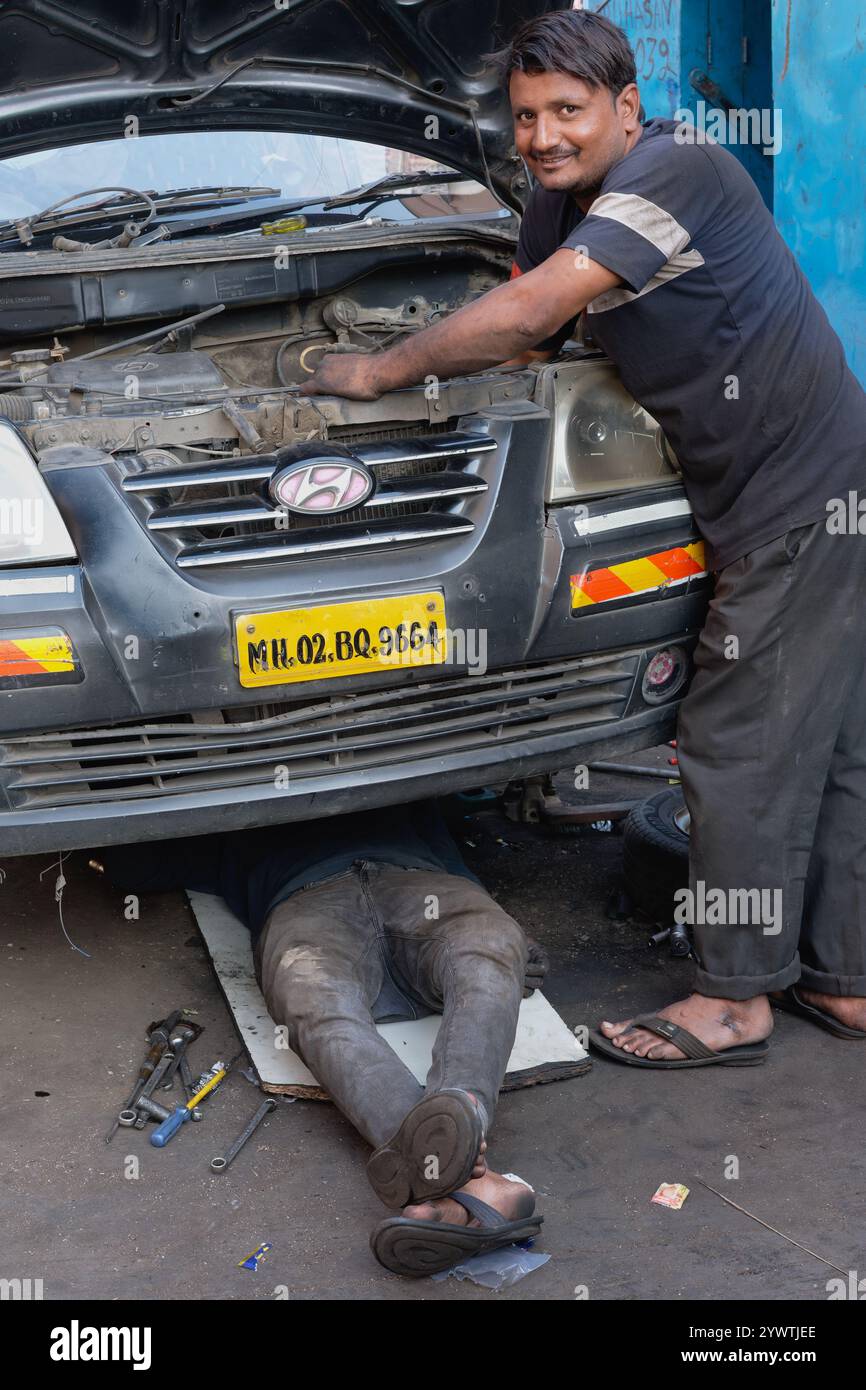 Zwei junge indische Autoreparaturmänner in Mumbai, Indien, arbeiten am Motor eines Autos, einer von ihnen arbeitet unter dem Fahrzeug Stockfoto