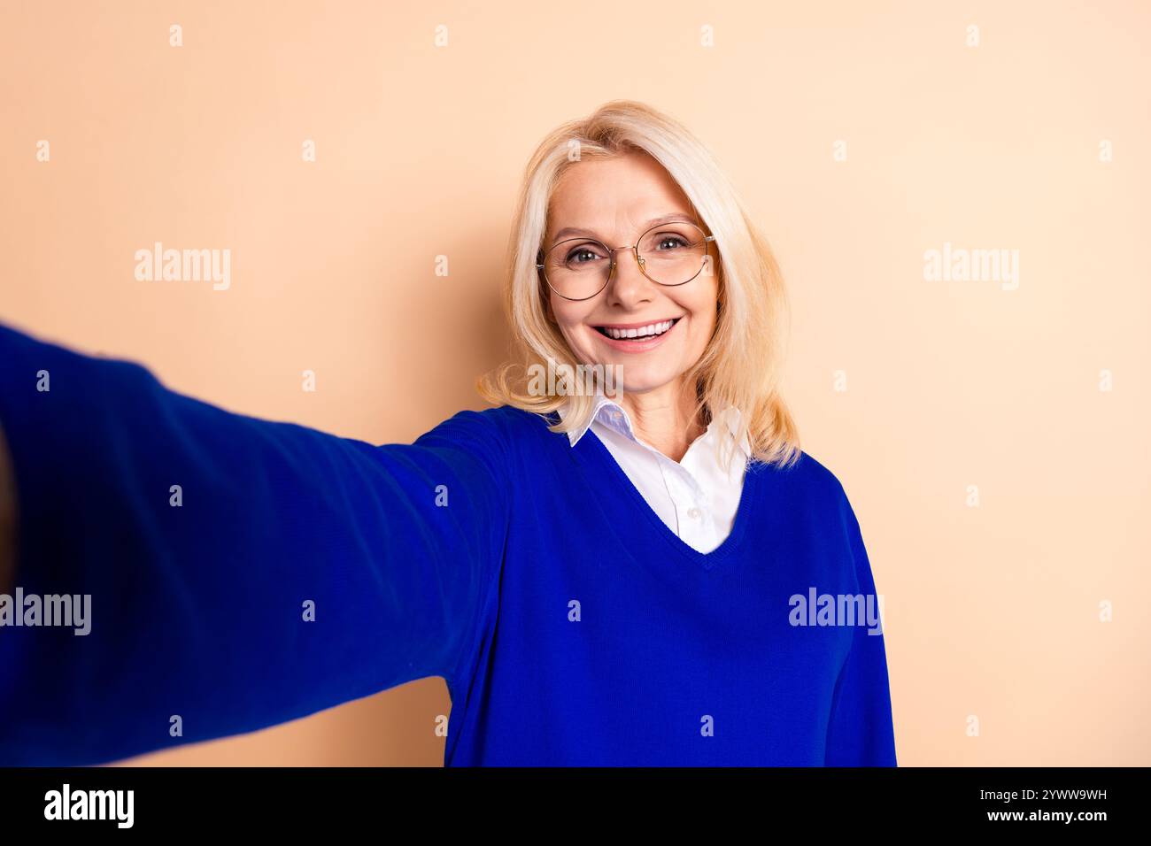 Elegante, reife Frau lächelt in blauem Pullover vor beigefarbenem Hintergrund und fängt professionelle und selbstbewusste Essenzen in lässiger Kleidung ein. Stockfoto