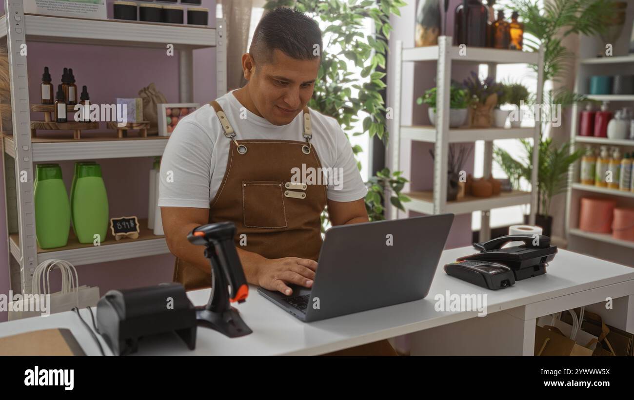 Junger Mann, der in einer Bäckerei arbeitet, einen Laptop benutzt, umgeben von Produkten und dekorativen Artikeln in Regalen, mit Pflanzen, die dem ein frisches Ambiente verleihen Stockfoto