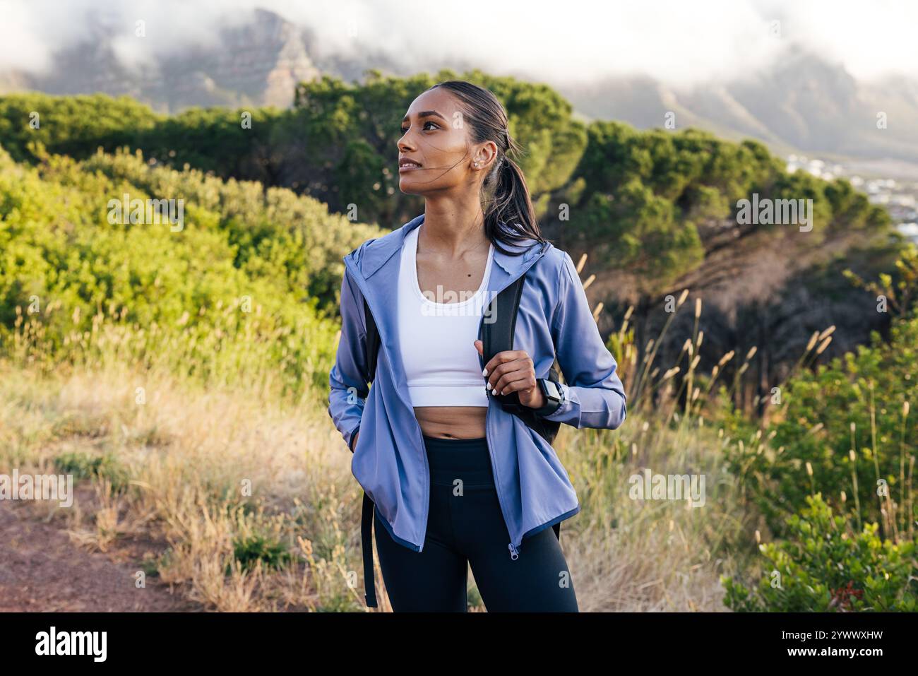 Wanderer in Sportswear, die wegschaut, während sie bei Sonnenuntergang gegen Berge mit Wolken steht. Läuferin, die eine Pause macht. Stockfoto