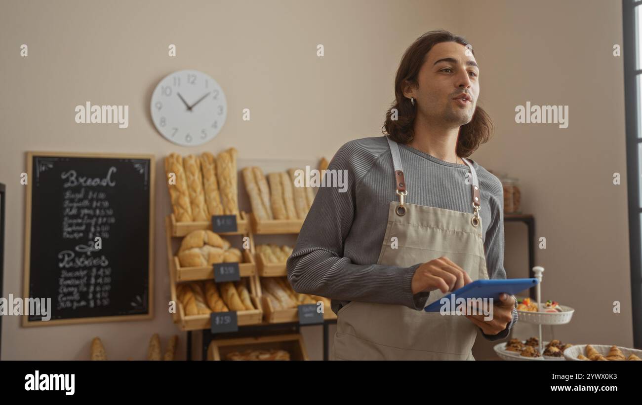 Junger hispanischer Mann mit langen Haaren und Schürze, der Tablette in einer Bäckerei hält, umgeben von Regalen mit Brot und Gebäck. Stockfoto