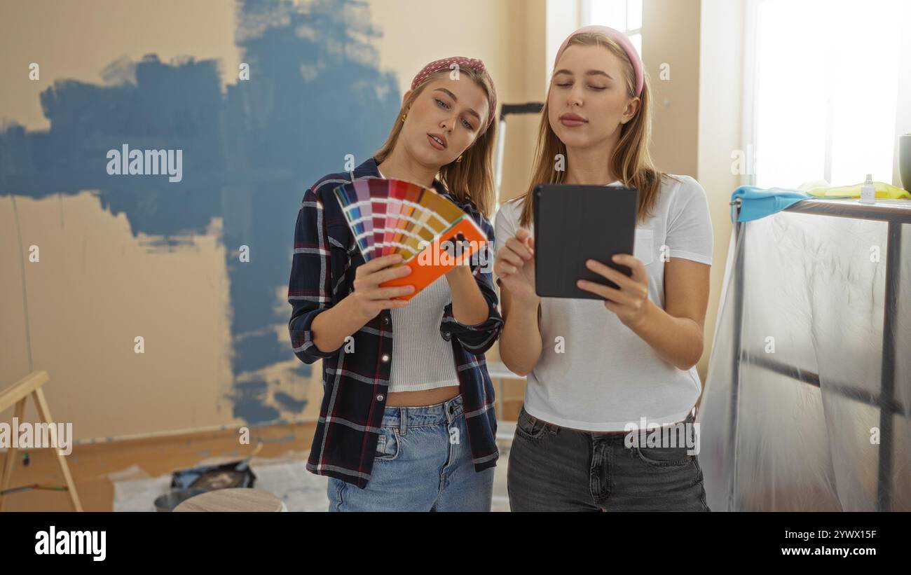 Frauen, die die Farbpalette gemeinsam in einem neuen Zuhause auswählen, zwei blonde Schwestern im Wohnzimmer, beide halten ein digitales Gerät in der Hand, bereiten sich auf die Zimmerrenovierung vor Stockfoto