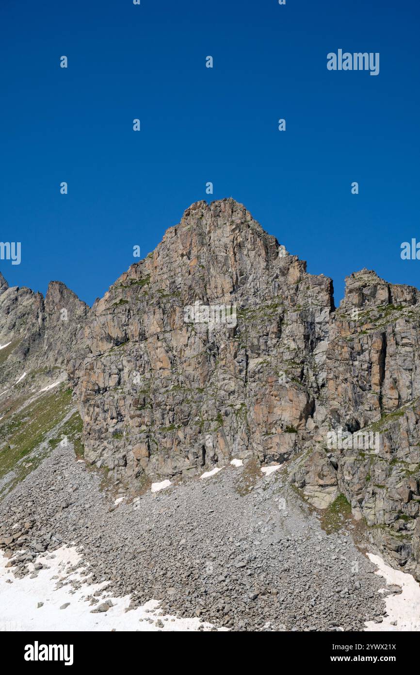 Hoch aufragender felsiger Gipfel in den italienischen Alpen mit zerklüfteten Klippen, Schneefelken und grüner Vegetation unter einem klaren blauen Himmel. Stockfoto