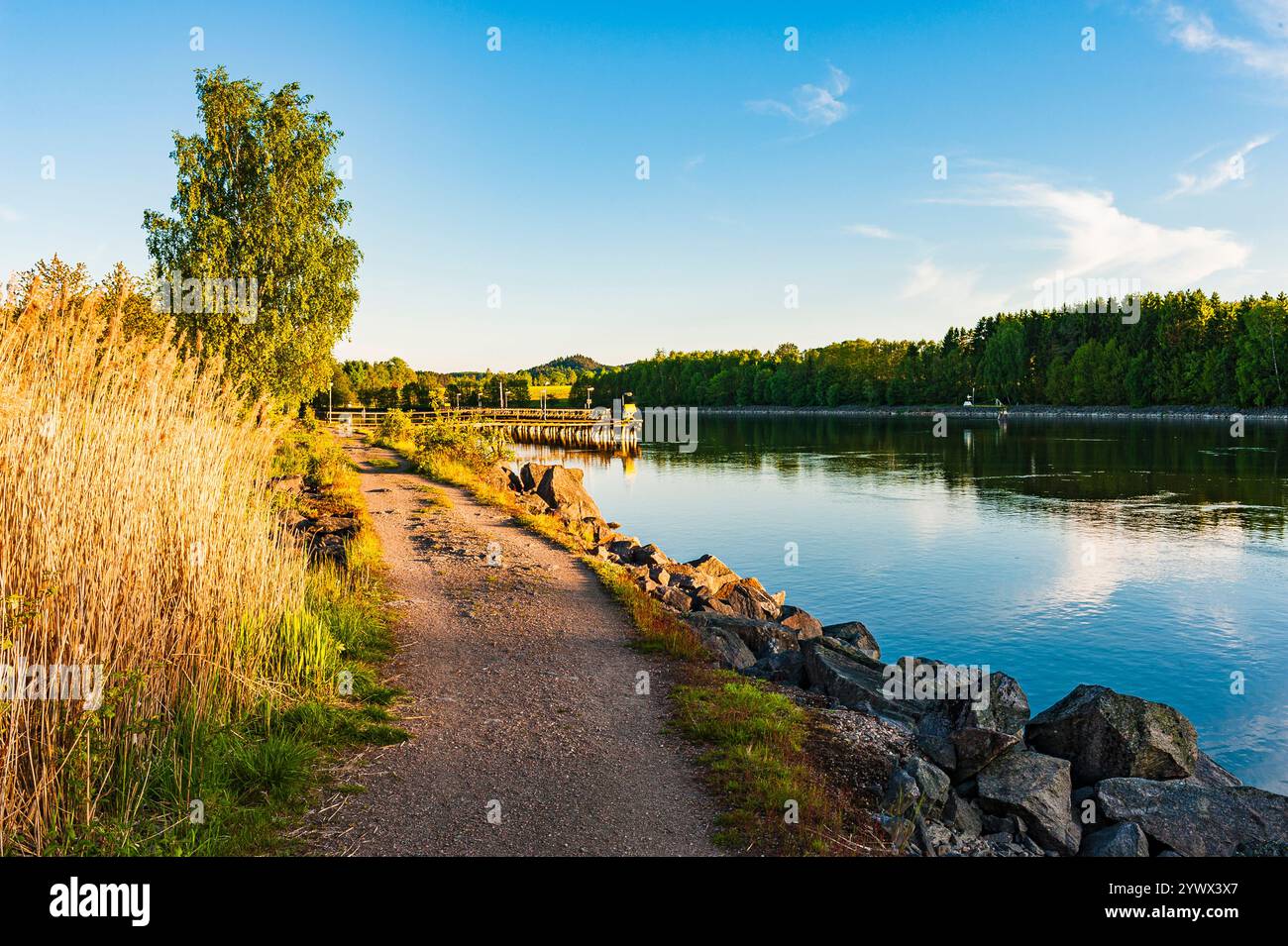 Ein friedlicher Flussweg, der von hohen Gras und Bäumen umgeben ist, lädt Besucher zum Erkunden ein. Ruhiges Wasser reflektiert den Sonnenuntergangshimmel und schafft einen ruhigen geldautomaten Stockfoto