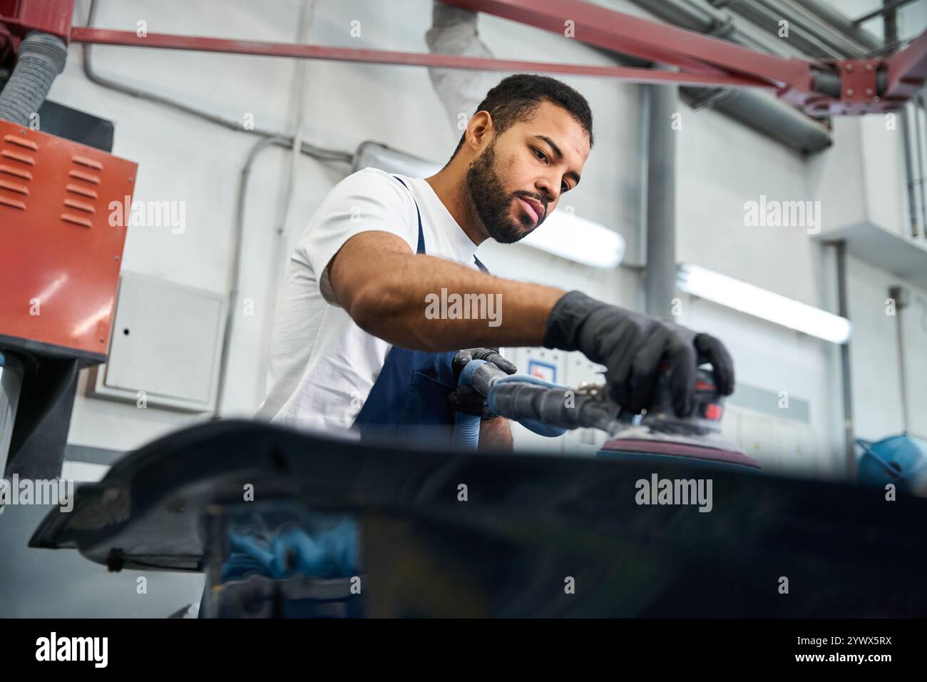 Ein gutaussehender Mechaniker verwendet ein Elektrowerkzeug, um ein Fahrzeugteil in einer belebten Garage zu perfektionieren. Stockfoto