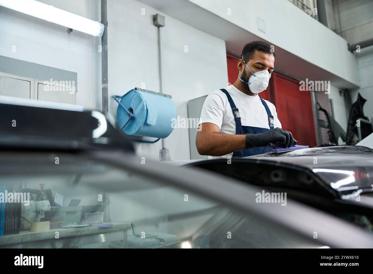 Gut aussehender junger Mechaniker in Arbeitskleidung inspiziert ein Fahrzeug in einer geschäftigen Autowerkstatt. Stockfoto