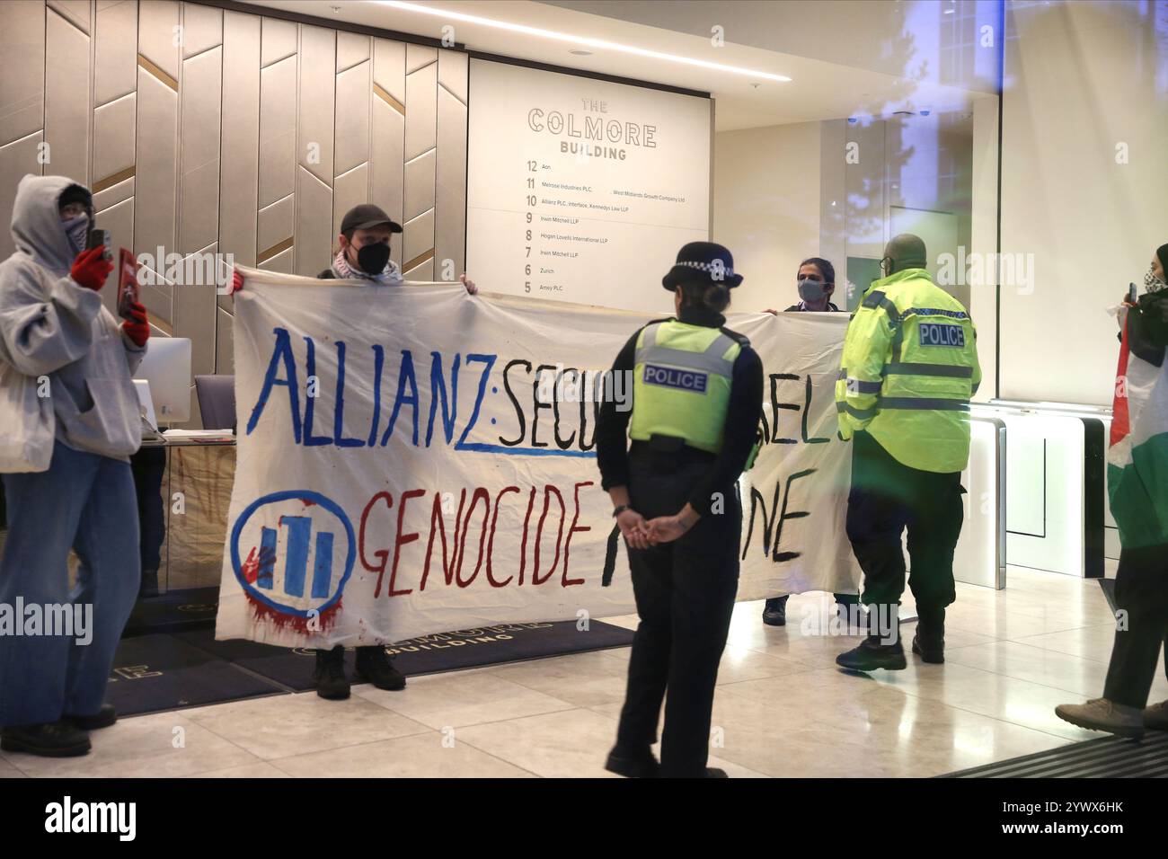 Bimingham, England, Großbritannien. Dezember 2024. Aktivisten protestieren im Empfangsbereich des Bürogebäudes mit einem Banner mit der Aufschrift „˜Allianz sichert Israels Völkermord-Maschine“ während der Demonstration, während Polizeibeamte anwesend sind und sie zum Verlassen bringen. Pro-palästinensische Aktivisten zielen auf die Versicherungsbüros der Allianz in ihrem Colmore Building in Birmingham, die den Empfangsbereich besetzen und draußen protestieren. Sie zielen auf die Allianz ab, weil sie das israelische Rüstungsunternehmen Elbit Systems versichern und in das israelische Rüstungsunternehmen Elbit Systems investieren und entschlossen sind, den Druck des direkten Handelns aufrecht zu erhalten, damit es zu einem großen Nachteil für wird Stockfoto