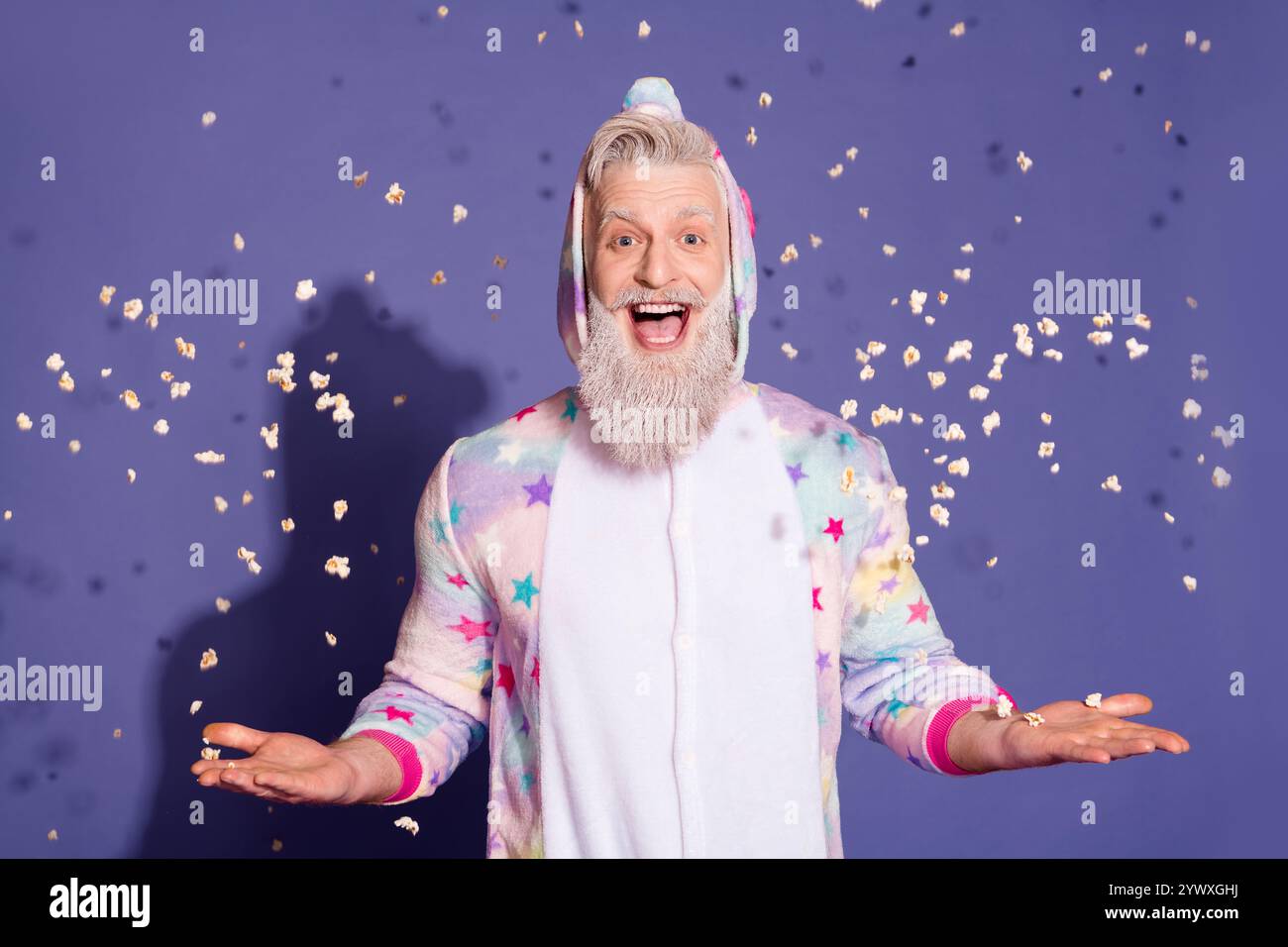 Fröhlicher Mann im farbenfrohen Einhorn-Pyjama, umgeben von fliegendem Popcorn auf lila Hintergrund Stockfoto