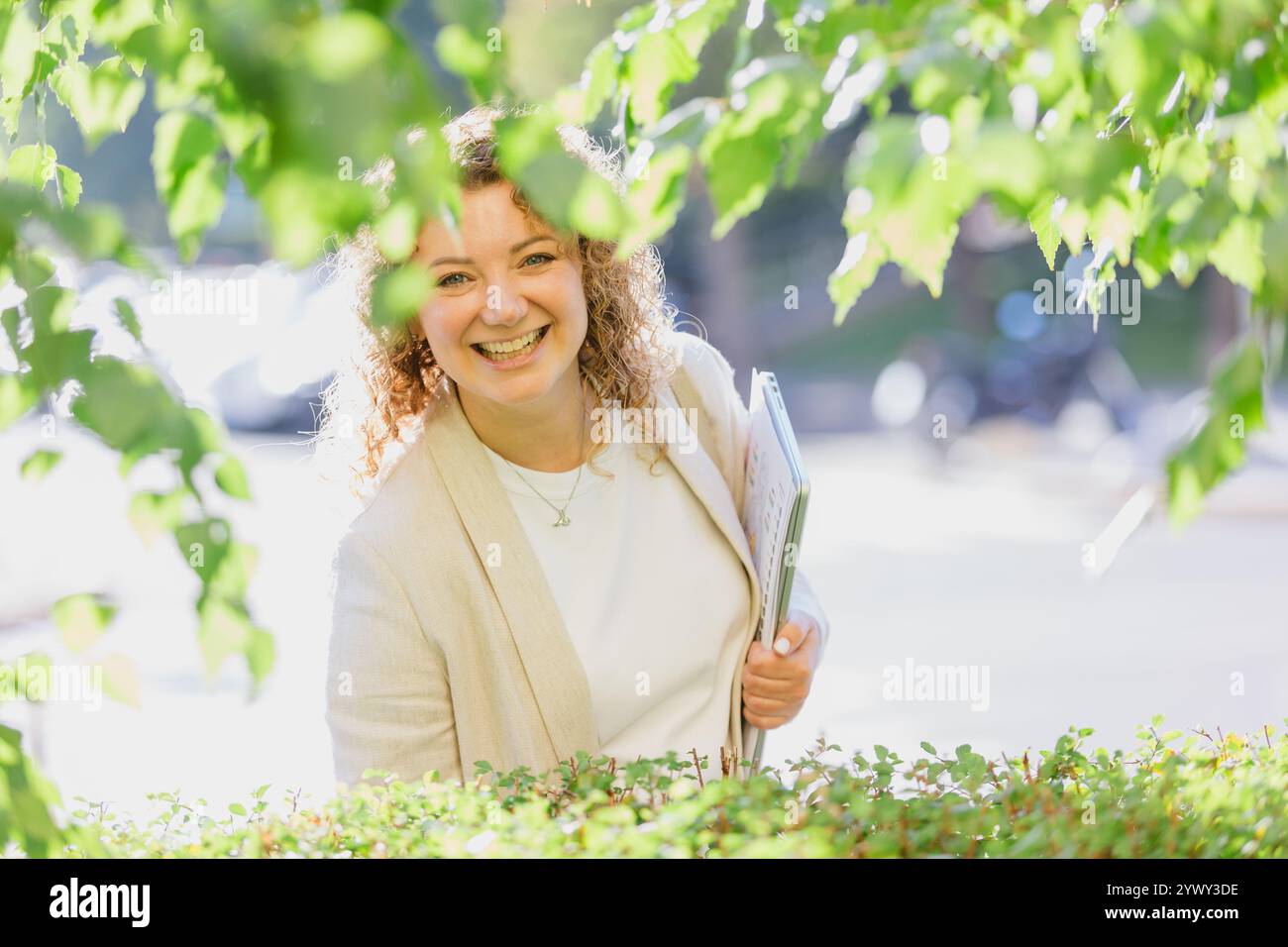 Professionelle Frau mit lockigen Haaren, gekleidet in einem weißen Hemd und einer beigen Jacke, steht draußen inmitten üppigen Grüns und hält Dokumente in der Hand Stockfoto