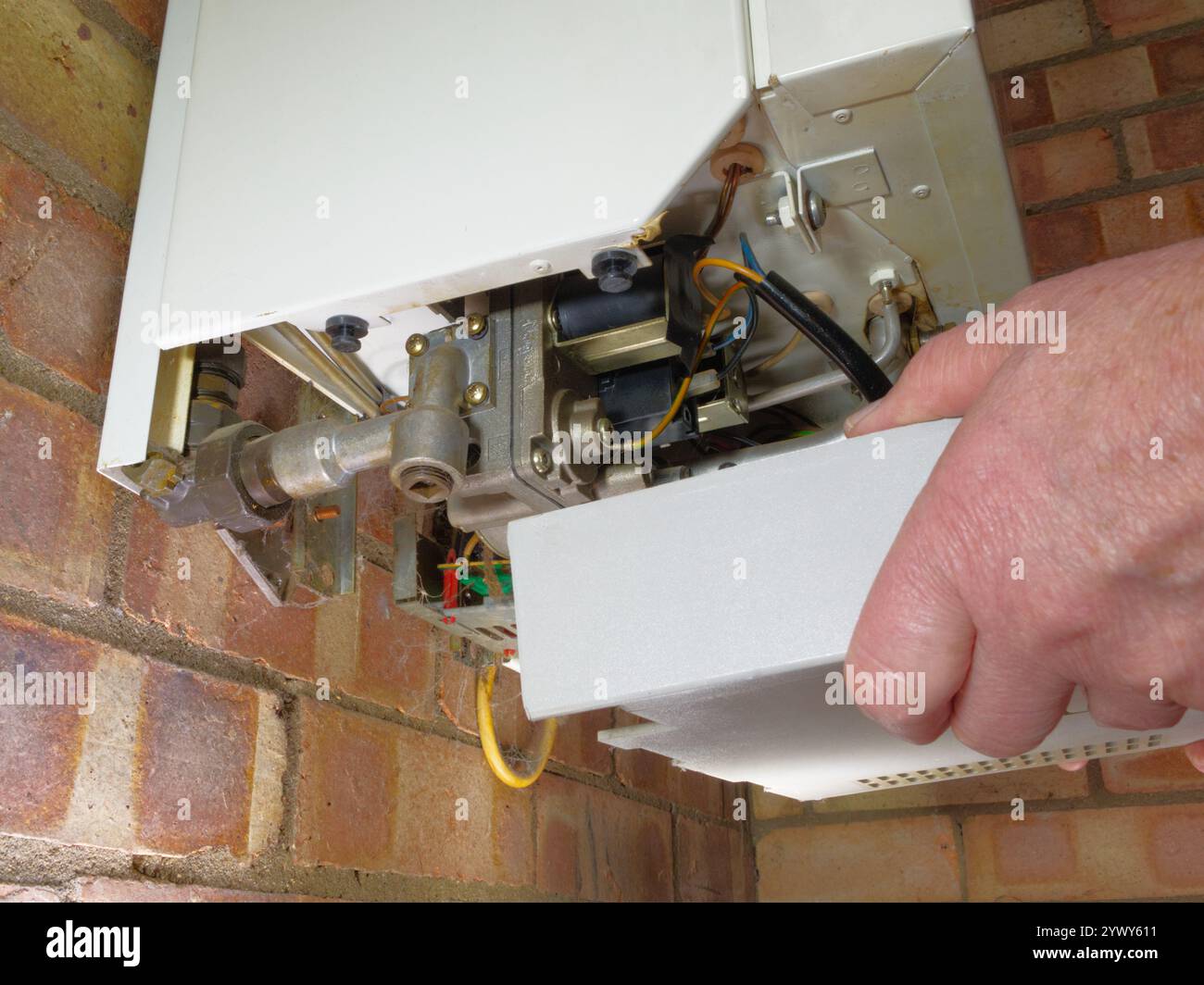 Nahaufnahme der Hände eines Mannes, der die Unterseite eines traditionellen Gaskessels für die Heizung und das Warmwasser hält, montiert auf einer inneren Ziegelwand. Stockfoto