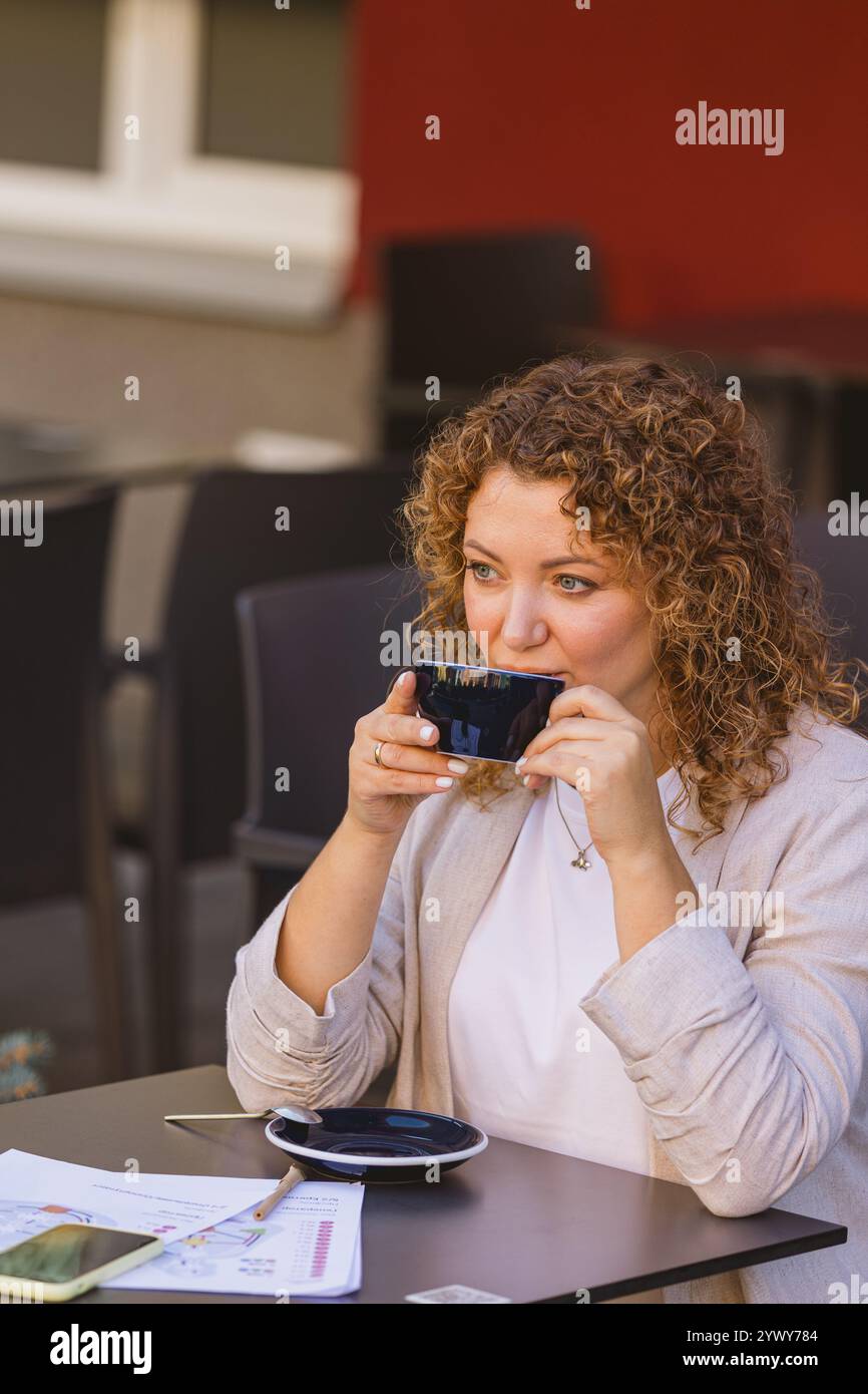 Eine Frau mit lockigem Haar genießt eine Tasse Kaffee, während sie in einem Café im Freien sitzt. Sie ist umgeben von Dokumenten und einem Smartphone, was eine Mischung anzeigt Stockfoto
