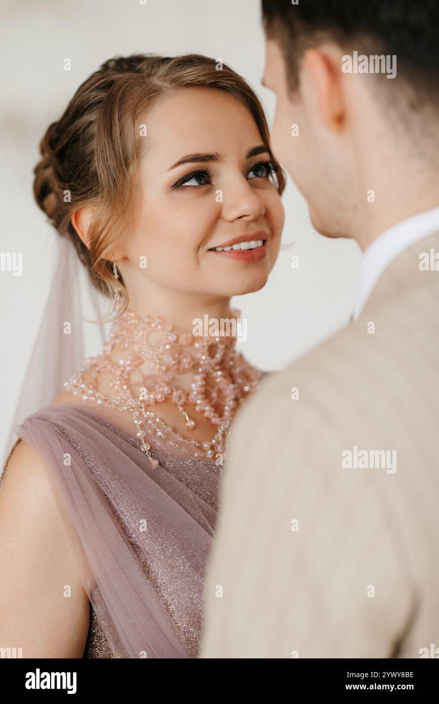 Bräutigam in beigem Anzug und Braut in lavendelfarbenem Hochzeitskleid in hellem Studio Stockfoto