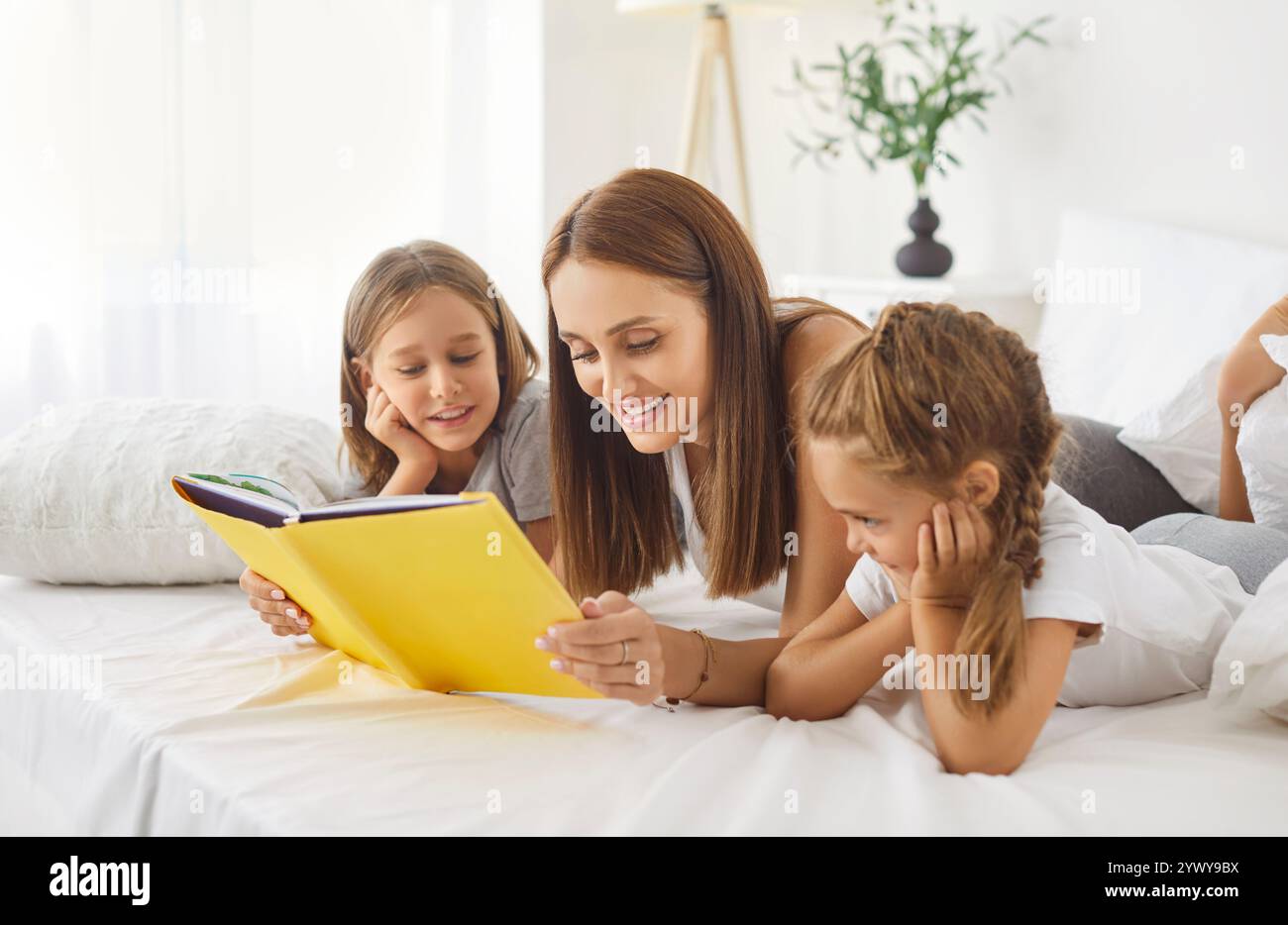 Lächelnde Mutter und ihre beiden Kinder liegen auf dem Bett, lesen zusammen Buch und genießen das Erzählen von Geschichten. Stockfoto