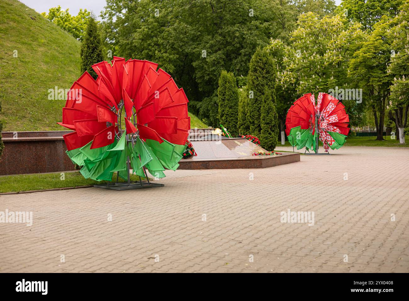 Denkmal mit ewiger Flamme umgeben von symbolischen roten und grünen Fahnen auf einem gepflasterten Platz in einem Park an einem bewölkten Frühlingstag Stockfoto