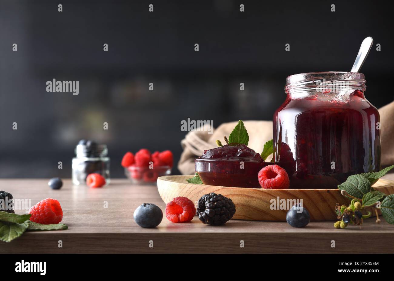 Waldfruchtmarmelade in Glasgläsern auf einer Holzbank mit Beeren in einer schwarzen Küche. Vorderansicht. Stockfoto