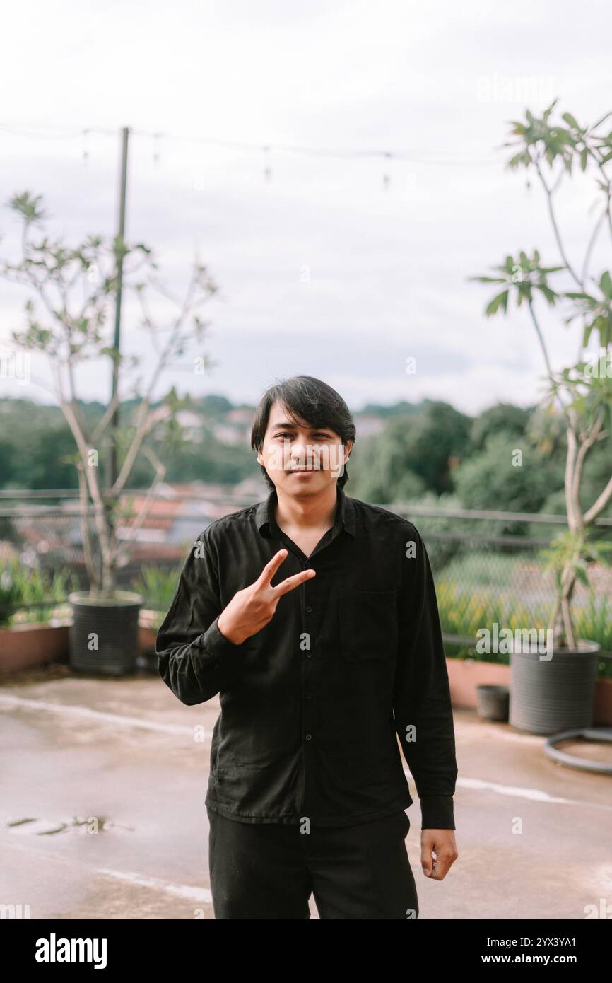 Ein südostasiatischer Mann, der mit den Fingern ein Friedenszeichen macht, das Harmonie und Positivität symbolisiert. Perfekt für Lifestyle, Frieden oder fröhlichen Ausdruck - der Stockfoto