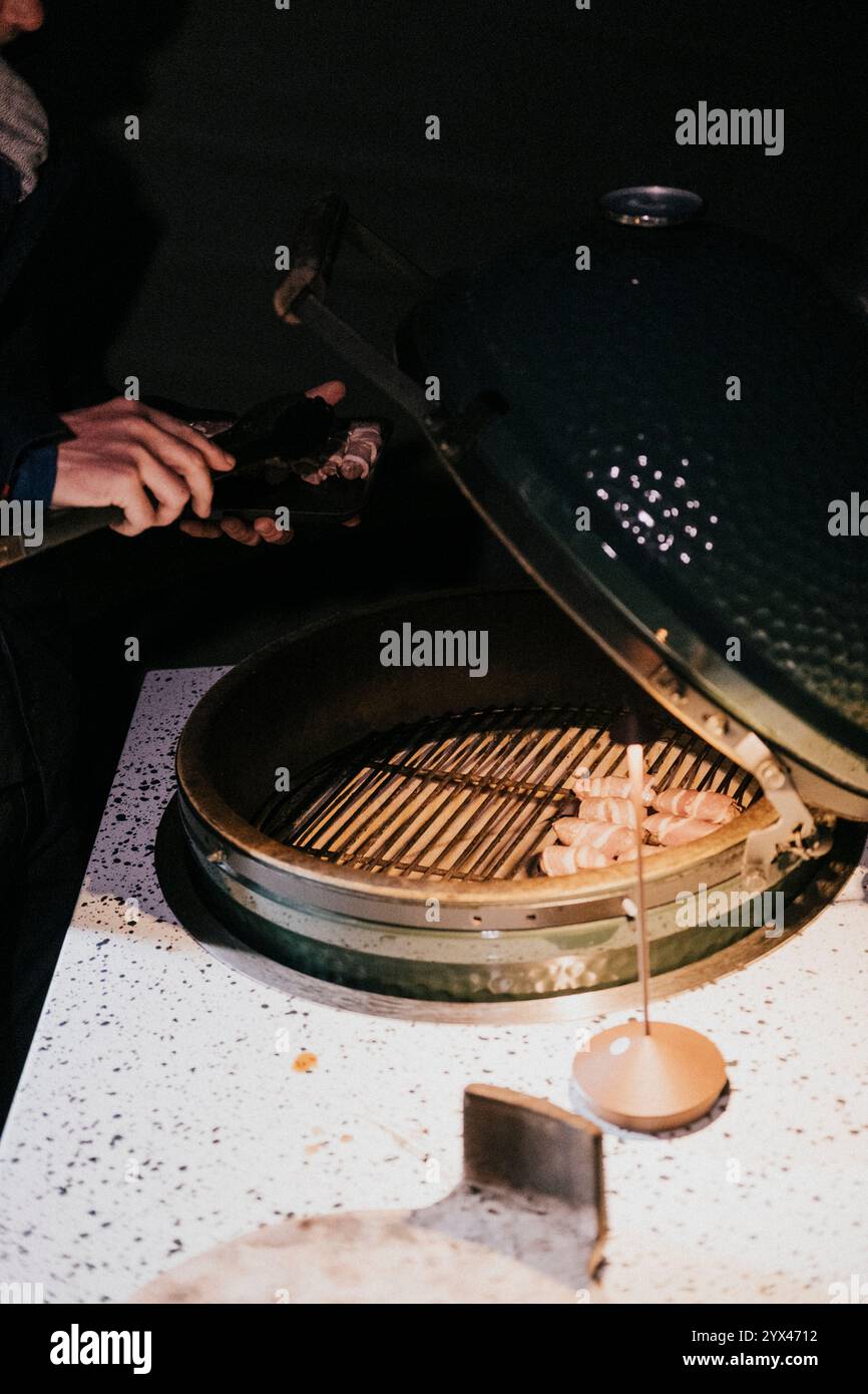 Eine Person kocht Essen auf einem Grill. Der Grill ist schwarz und hat einen Deckel. Die Person hält eine Flasche und ein Messer Stockfoto